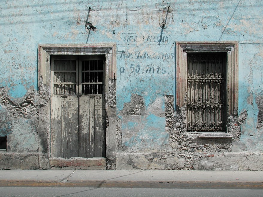 A worn-down wooden door lies framed by a crumbling building façade in Mérida. Traces of faded lettering remain where the paint has not flaked away.