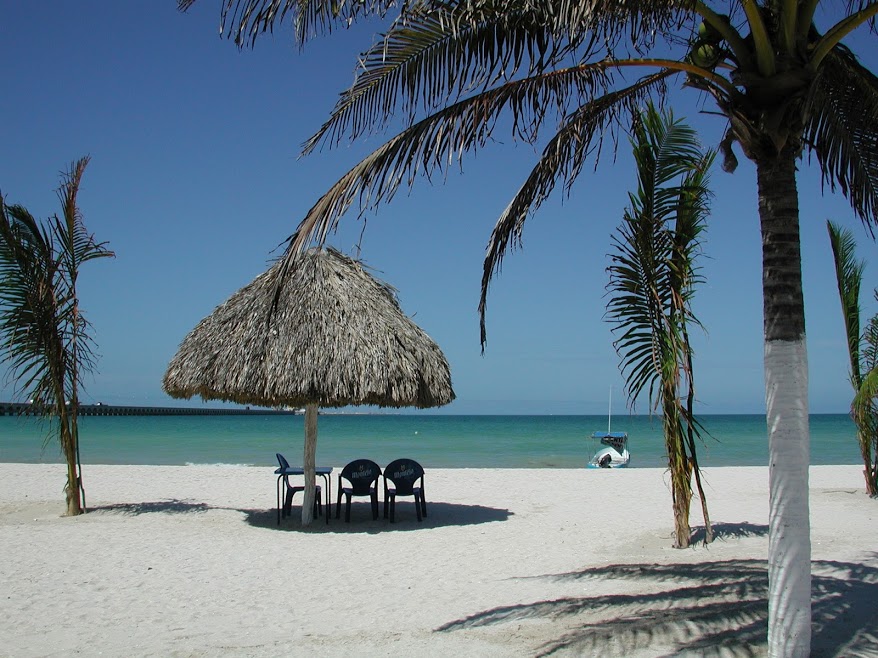 Three beach chairs sit in the shade of a palm-thatched palapa on the beach overlooking the ocean. A small 'lancha' boat is pulled up on the beach. On the left, Progreso's long pier extends over the water towards the horizon.