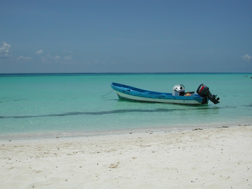 A small 'lancha' boat floats in the crystal-clear blue waters of the Caribbean, moored a few metres offshore a white sandy beach.