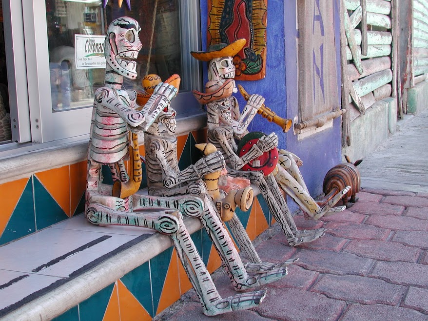 Four small hand-carved wooden skeleton toys playing musical instruments and wearing sombreros sit on the step of a storefront with their feet on the sidewalk. A small wooden armadillo wanders by.