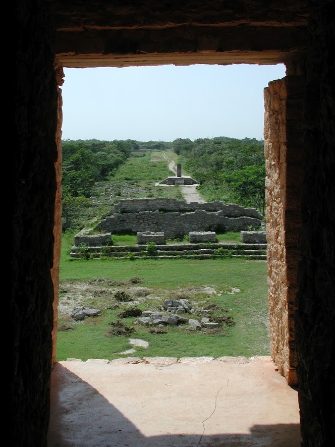 View framed by the doorway of the of Templo de las Siete Muñecas looking out over the ruins of a stone building and four-sized stone stela on a raised platform. A path leads past the ruins, through the low jungle, and towards the horizon.