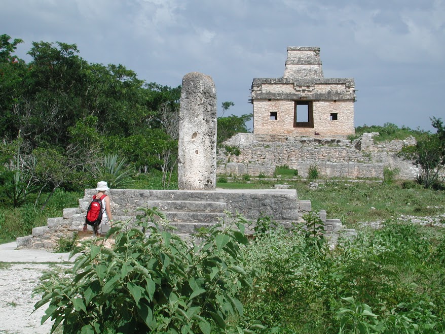 View of the Templo de las Siete Muñecas from the path. In the foreground, a hiker walks toward a large worn stela on a raised platform.